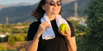 Mulher sorrindo com toalha e maçã verde em área externa.
