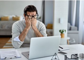 Homem de óculos sentado em frente ao laptop com as mãos no rosto, aparentando estresse ou cansaço durante o trabalho.