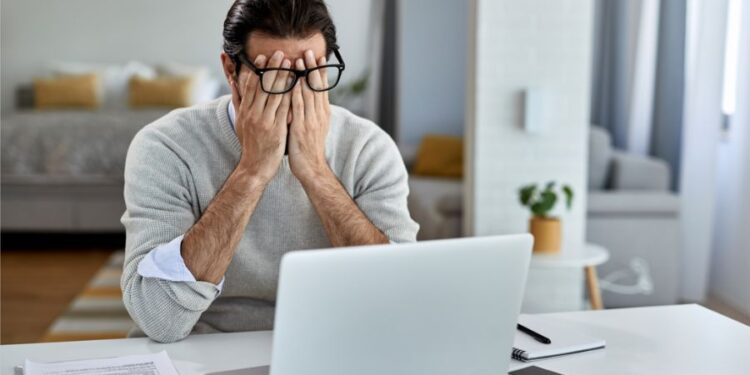 Homem de óculos sentado em frente ao laptop com as mãos no rosto, aparentando estresse ou cansaço durante o trabalho.
