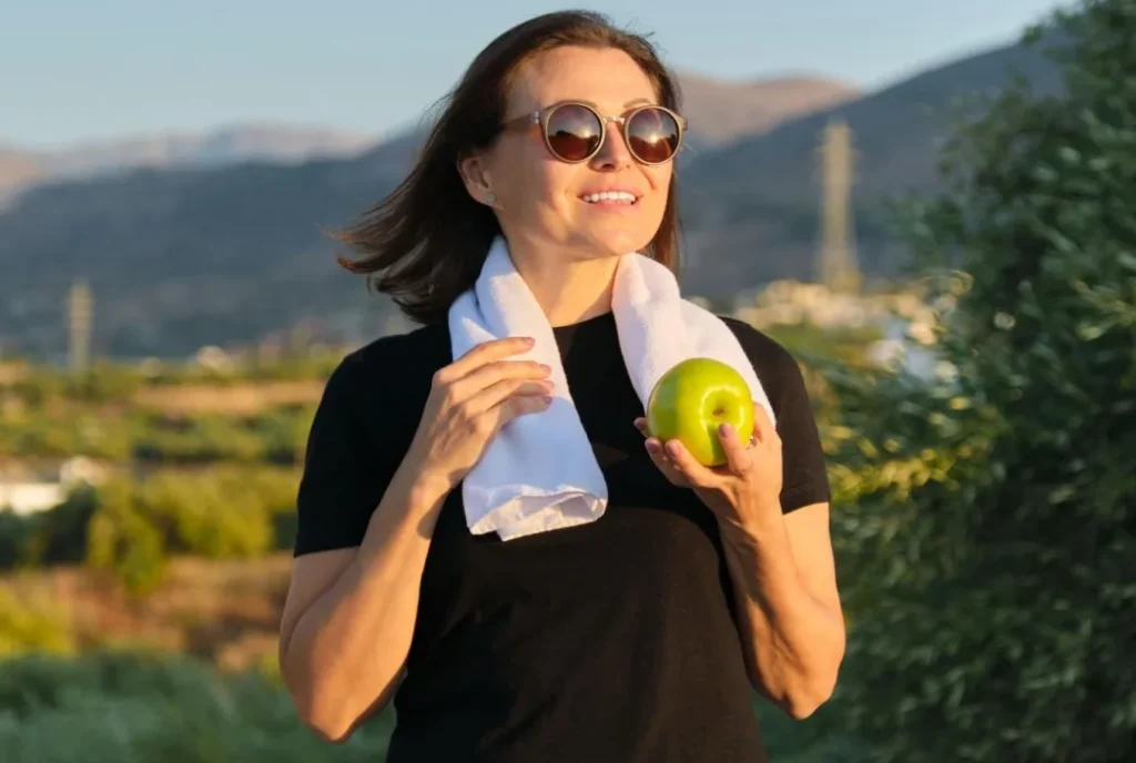 Mulher sorrindo com toalha e maçã verde em área externa.