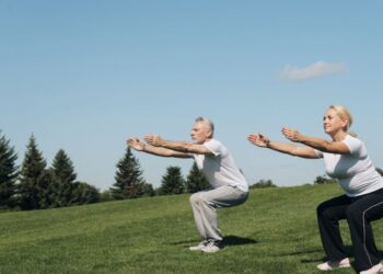 Casal de idosos praticando exercício físico ao ar livre, fazendo agachamento em um campo gramado ensolarado