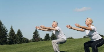 Casal de idosos praticando exercício físico ao ar livre, fazendo agachamento em um campo gramado ensolarado