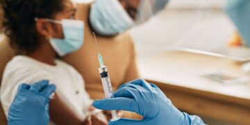 Criança usando máscara sendo preparada para vacinação por profissional de saúde