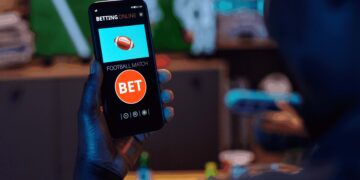 Foto de homem, visto de costas, segurando um celular, que exibe um aplicativo de apostas.