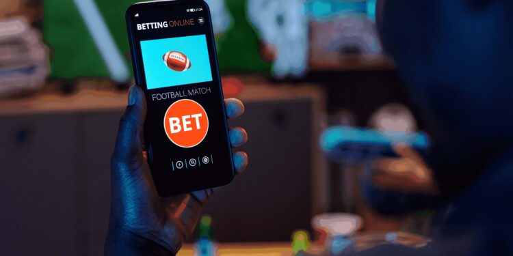 Foto de homem, visto de costas, segurando um celular, que exibe um aplicativo de apostas.